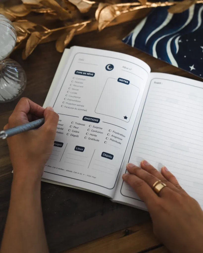 Mains tenant le Journal de Rêves ouvert sur une table en bois, montrant les sections guidées : type de rêve, émotions, personnages, lieux et objets sur la page de gauche, espace de notes ligné sur la page de droite.