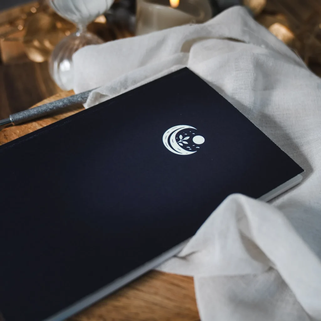 Couverture rigide noire du Journal de Rêves avec logo croissant de lune, plante et étoiles en blanc, posée sur table en bois avec ambiance cosy