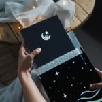 Mains tenant le Journal de Rêves dans son packaging avec couverture noire ornée du logo croissant de lune et plante, dans une ambiance chaleureuse avec guirlandes lumineuses